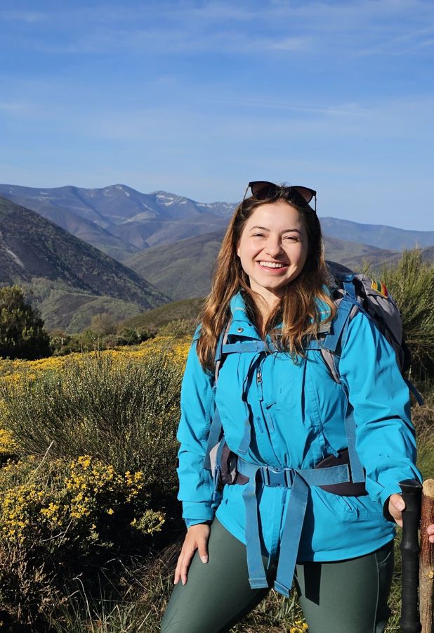 Elisa sorrindo, de jaqueta azul e mochila, com montanhas ao fundo