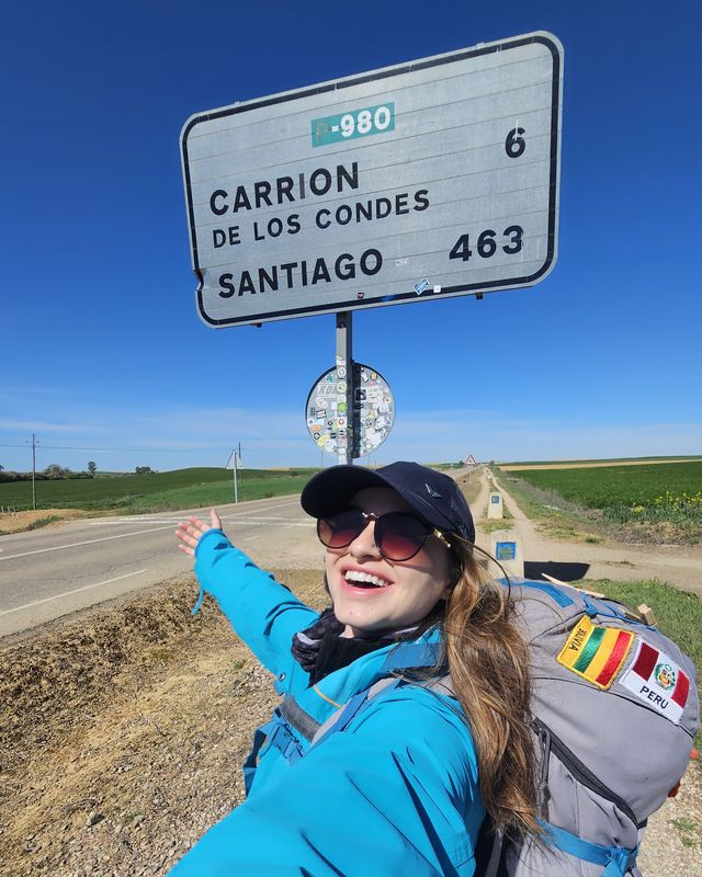 Elisa de mochila apontando para a placa: Carrión de los Condes 6, Santiago 463